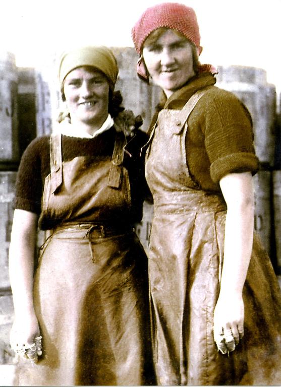 Archive image of two women wearing leather aprons. The woman on the left is wearing a yellow bandana headscarf and the woman on the right is wearing a one in red.
