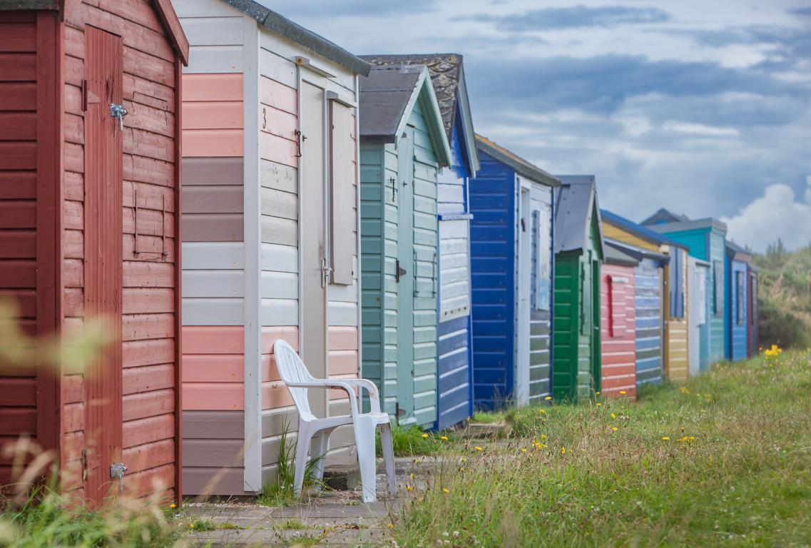 Row of pastel coloured beach huts sit on a grassy verge.