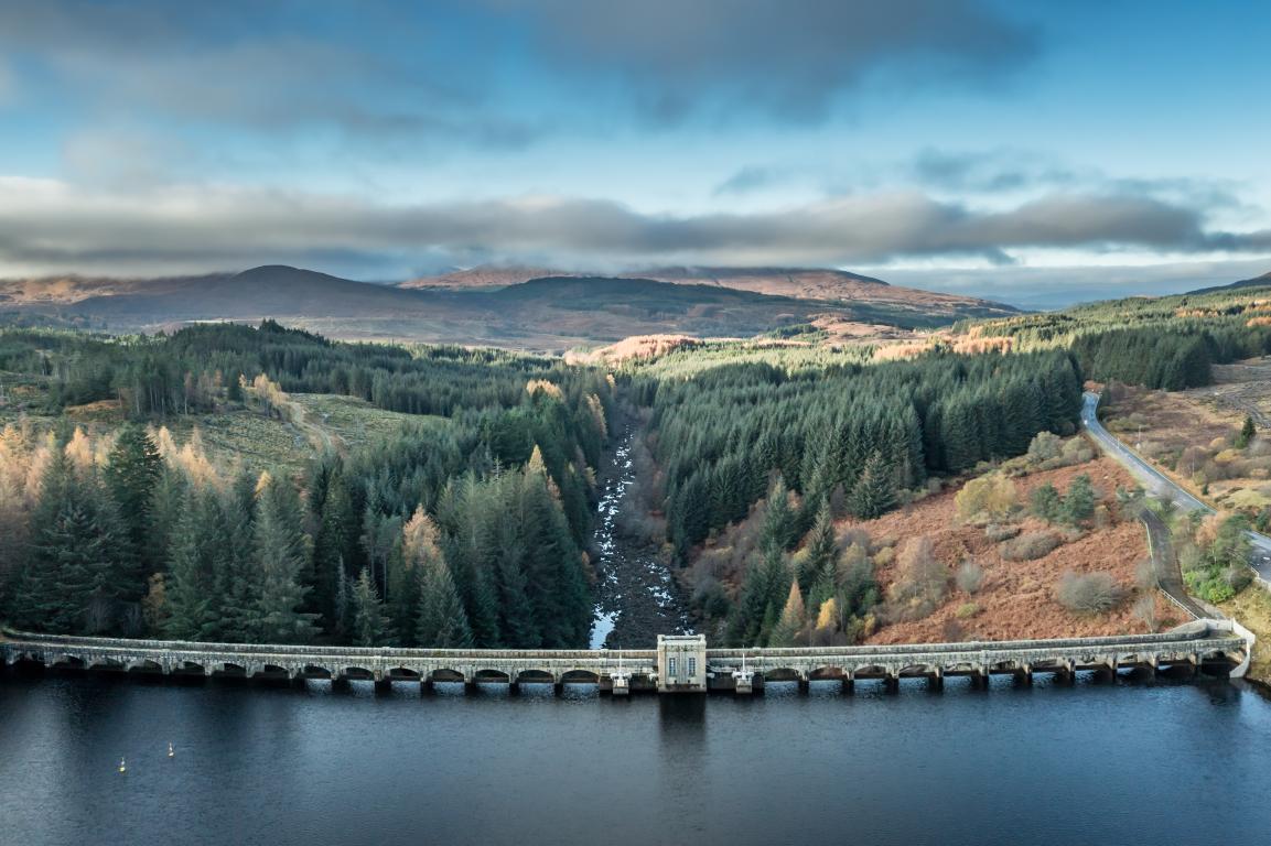 Bridge above dam overlooks a rocky loch lined with pine trees and heading towards a hilly backdrop.