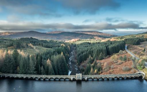 Bridge above dam overlooks a rocky loch lined with pine trees and heading towards a hilly backdrop.