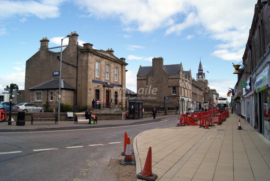Looking east onto Nairn High Street. Large buildings line the high street, with a spire visible in the background of the image