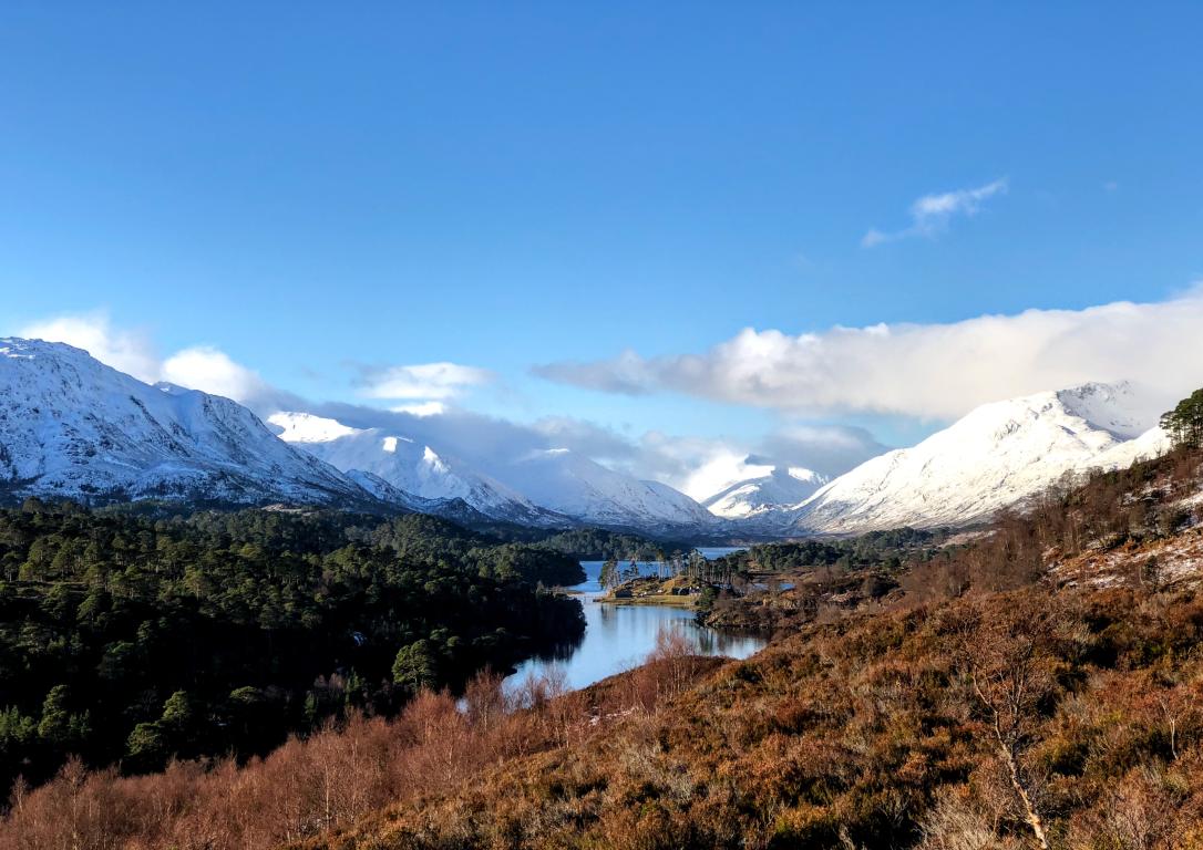 Glen Affric in winter with mountains in the top half of the image covered in snow. Clouds brush the top of the mountain peaks under a clear blue sky.