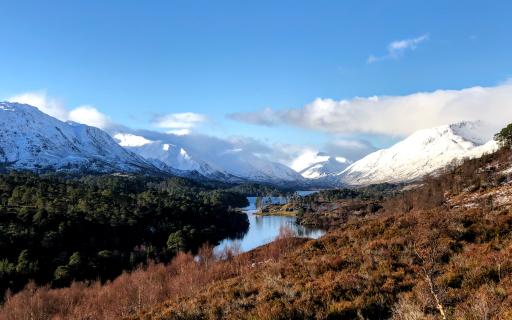 Glen Affric in winter with mountains in the top half of the image covered in snow. Clouds brush the top of the mountain peaks under a clear blue sky.