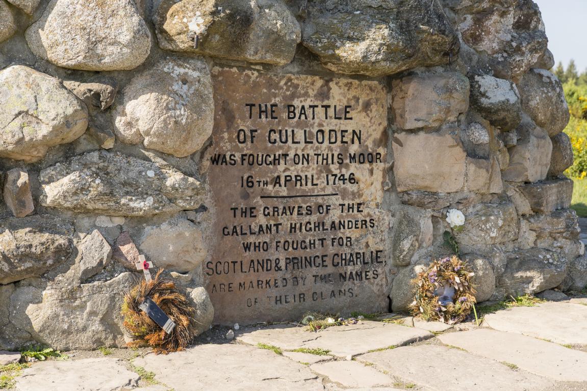 An engraving on Culloden Memorial which reads "The Battle of Culloden was fought on this moor 16th April 1746. The graves of gallant Highlanders who fought for Scotland & Prince Charlie, are marked by the names of their clans." Two brown circular wreaths are placed either side of the engraved panel