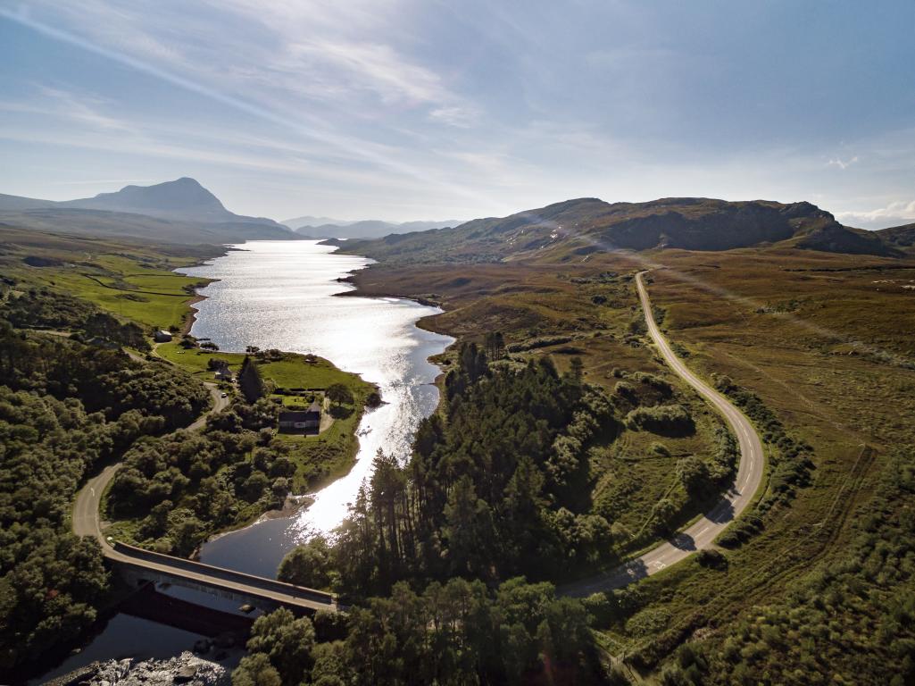 Loch Hope, Near Tongue flanked by a softly sloping green landscape. A winding road curves across the loch from the right and continues on to the left hand side of the loch.