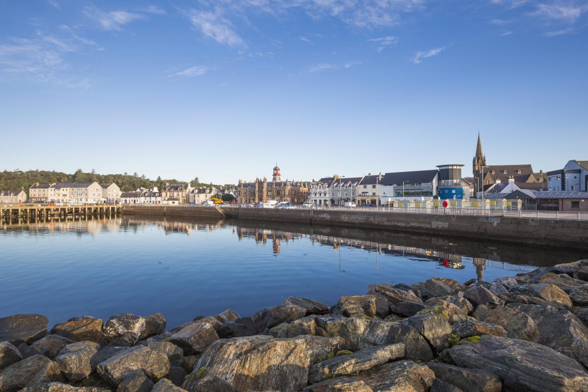 Stornoway Harbour, Isle of Lewis. Stones are piled across the harbour edge in the bottom 25% of the image. Colourful shop front line the harbour in the center of the image.