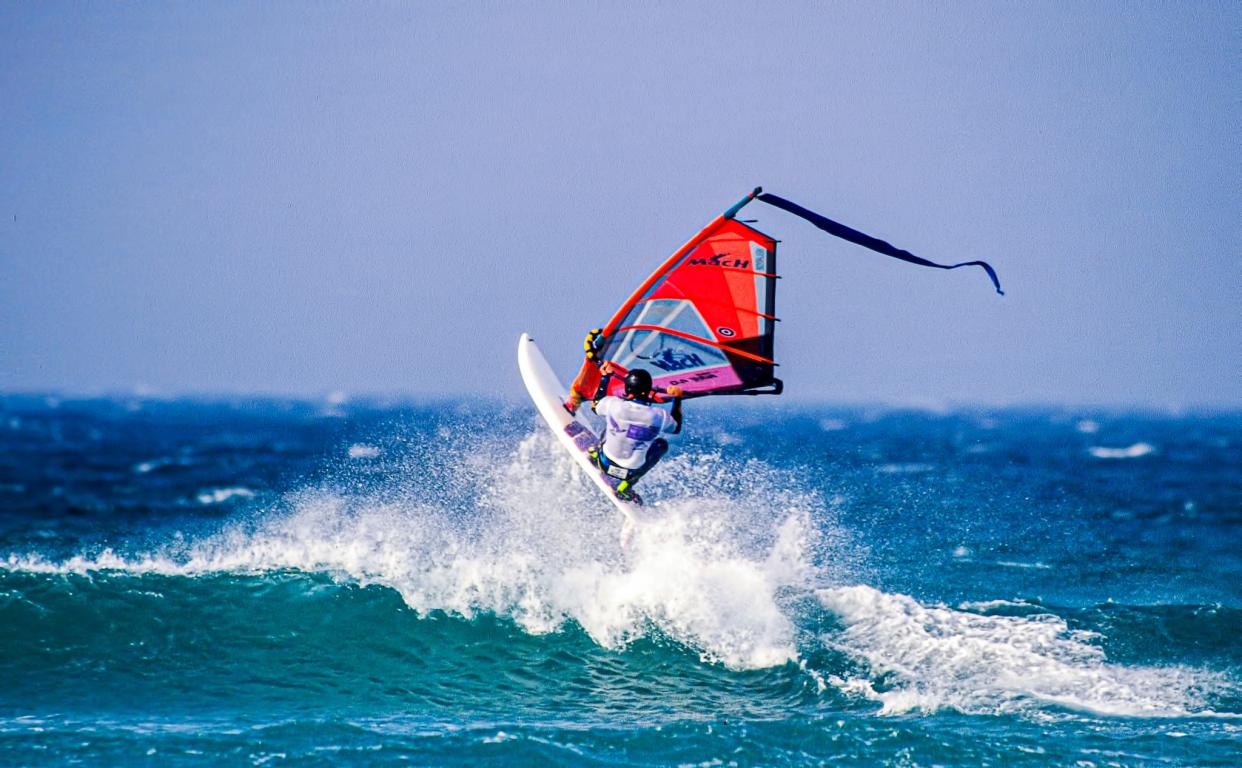 Windsurfer at top of wave with spray and blue sea lapping at the board.