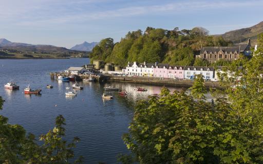 A view over Portree harbour, Isle of Skye. Colourful buildings line the shorefront. Sea vessels of varying sizes can be seen on the water in front of the pier. Green-leaved trees can be seen both in the distance and the foreground of the image.