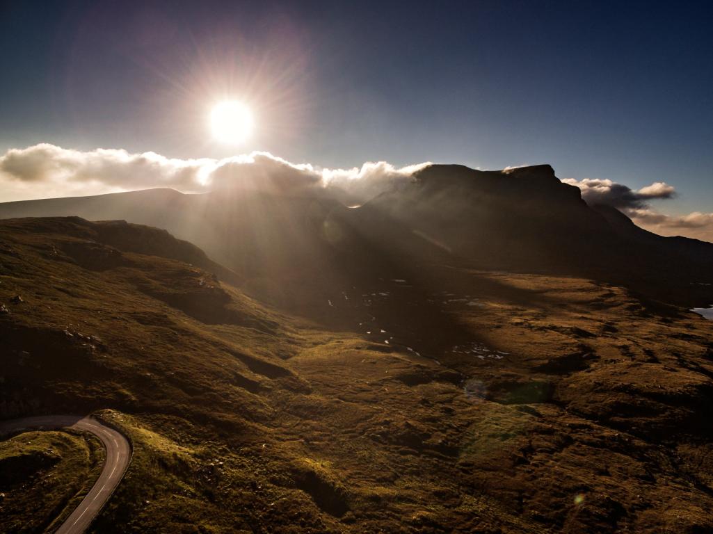 The outlines of mountains and a valley at sunset. The sun shines bright above a layer of cloud. Sheep can be seen roaming a field in the distance.