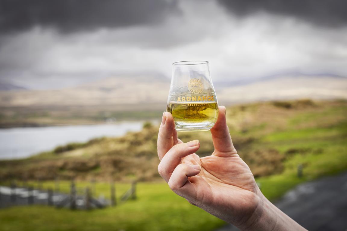 A small glass about half full with whisky is being raised by a hand in the foreground of the image.