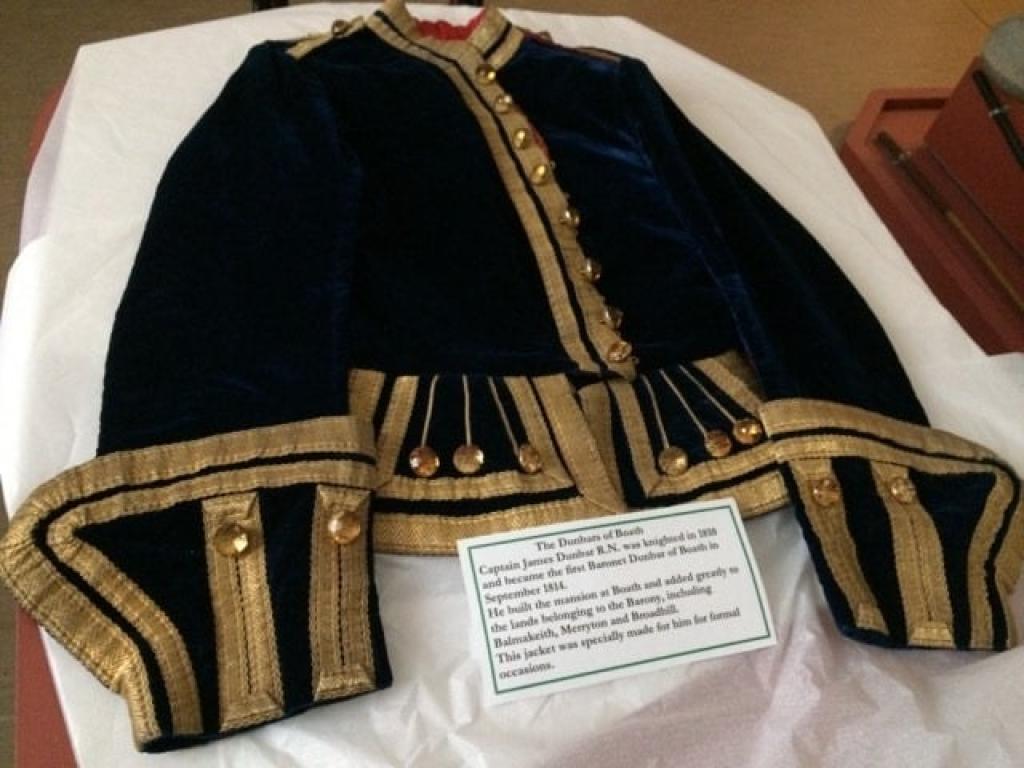 A large black men's jacket with gold buttons and gold lining sits atop a white backdrop. An information panel sits at the bottom of the jacket. The panel reads "The Dunbars of Booth. Captain James Dunbar R.N. was knighted in 1809 and became the first Baronet Dunbar of Booth in September 1814. He built the mansion at Booth and added greatly to the lands belonging to the Barony, including Balmakeith, Merryton and Broadhill. The jacket was specifically made for him for formal occassions."