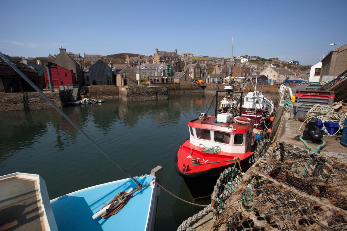 The harbour at Stromness. A boat, painted red can be seen in the foreground of the image. A boat painted blue is moored in front of the red boat.