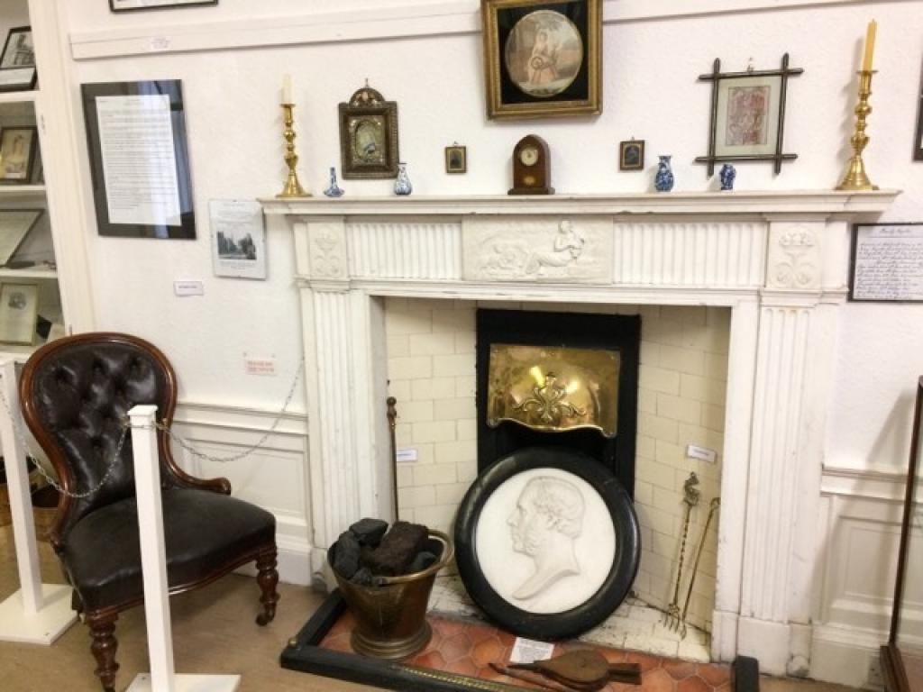 A white fireplace in front of which are artefacts from the history of Nairn. This includes a brown, armless chair on the left hand side of the image and a carving of the side profile of a man in the bottom center of the image