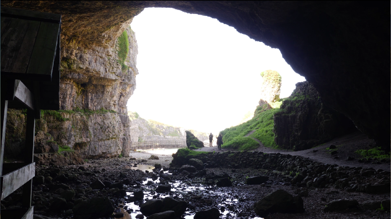 The entrance to a cave as seen from the inside. A small stream covered in rocks of varying sizes can be seen in the bottom half of the image. Two people can be seen at the entrance path to the cave.