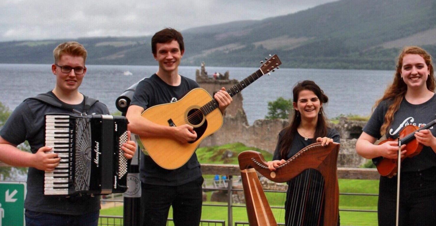 Five musicians gather outside Urquhart Castle. Each is wearing a black shirt which reads 'Fèis Rois'