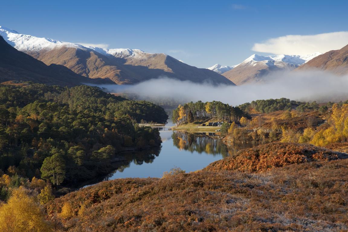 Looking across Glen Affric. Low mist covers the bottom of snow tipped mountains. Trees with leaves of green, yellow and orange line the glen.