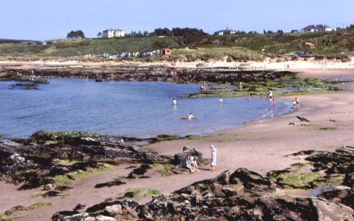 A sandy beach lined with rocky outcrops, with visitors scattered across the beach on the right hand side of the image