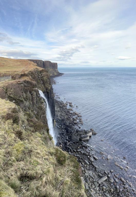 A steep cliff face dropping into the clear blue sea at Kilt Rock Point. A waterfall can be seen cascading down the cliff face in the first half of the image