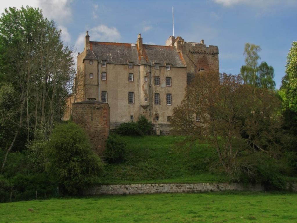 Kilravock Castle, a large light brown building with a rusting grey roof, stands on grassy knoll with two trees in the foreground.