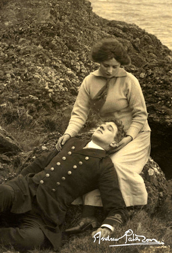 A woman with short brown hair in a collared shirt holding a man, unconscious, with short brown hair in a black vest jacket