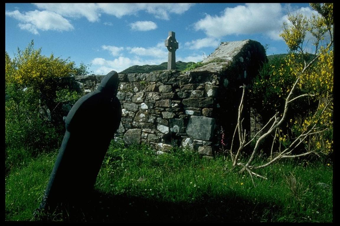 View of the exterior stone wall of St. Finnan's Chapel with a large tilted gravestone in the foreground. A large stone cross in the distance peers above the stone wall.
