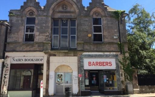 A large grey stone building with three large windows. Two shops - Nairn Bookshop, on the left, and a barber shop, on the right, are housed in the building