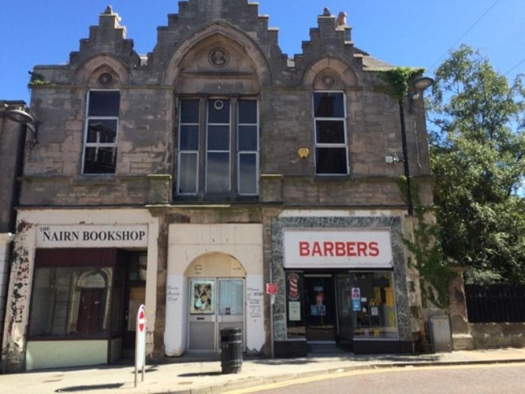 A large grey stone building with three large windows. Two shops - Nairn Bookshop, on the left, and a barber shop, on the right, are housed in the building
