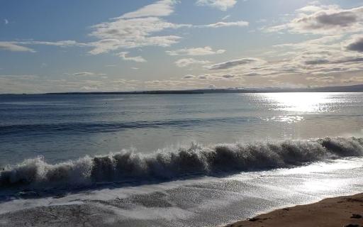 Rosemarkie sandy beach with wave coming in. The top 25% of the image features a light blue sky and sparse white clouds