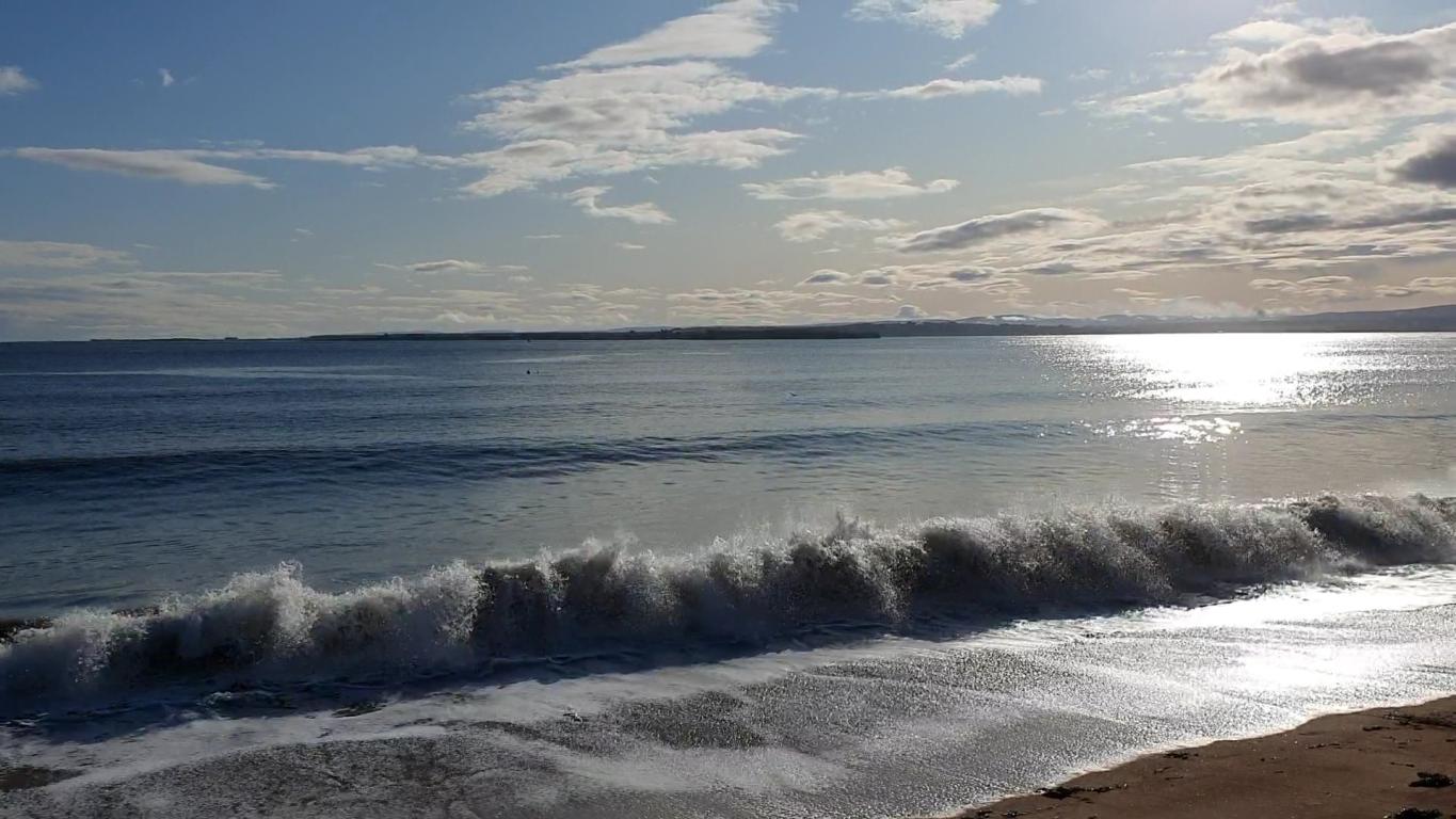 Rosemarkie sandy beach with wave coming in. The top 25% of the image features a light blue sky and sparse white clouds