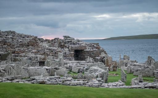 The ruins of a Scottish Iron Age stone building - a broch - stand on a grassy field under a cloudy sky. The sea and an outline of land can be seen in the distance behind the broch.