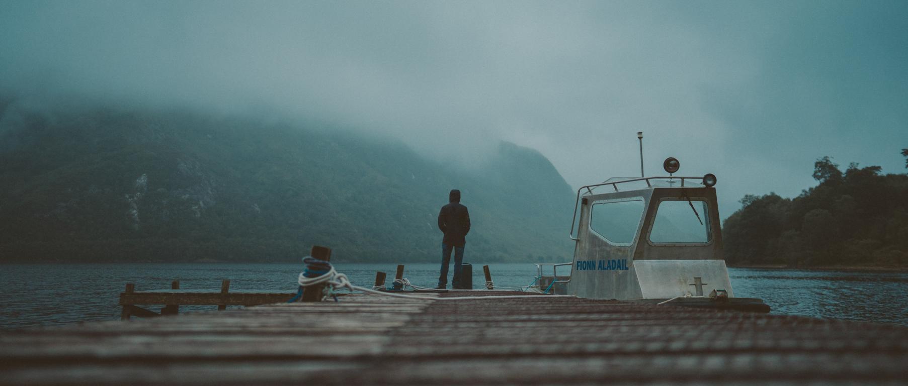 Man stands on pier with boat beside him looking out to water which is heavily covered in fog.
