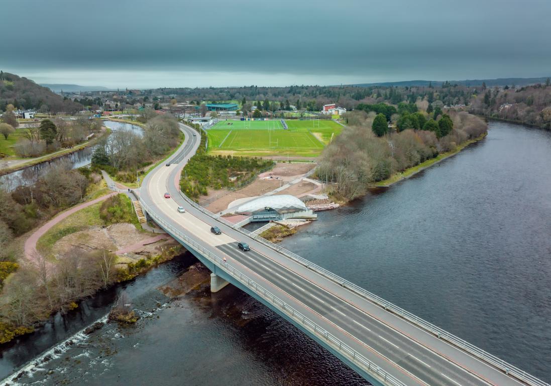 A silver oblong-shaped building, named the Hydro Ness, as seen from the air. A road bridge over the River Ness with 5 cars sweeps through just off-centre, on the left, of the image.