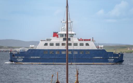 A blue and white ferry, the MV Leirna, is sailing in the centre of the image. A small wooden canoe boat with a large mast in buoyed in the forefront, visible in the bottom half of the image.