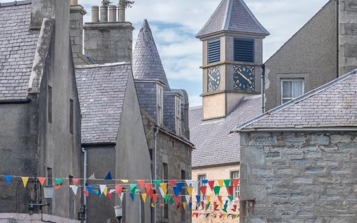 Grey rooftops and buildings with colourful bunting strung between them and town clock in background.