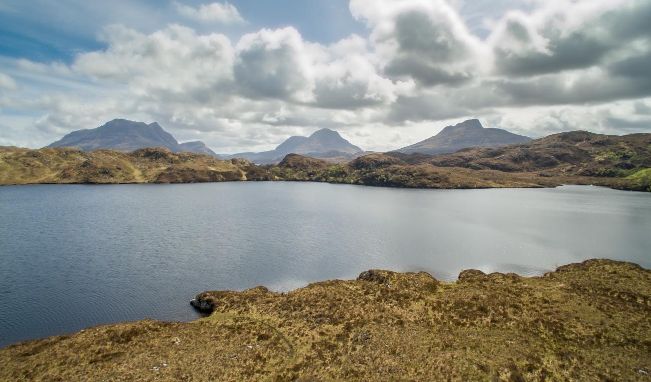 A light green-earthy brown landscape lines a body of still blue water. The jagged outlines of three mountains can be seen in the background under a cloudy sky.