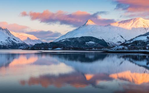 The sun setting over Glencoe. The mountain peaks are covered in heavy snow. The mountains and pink clouds are reflected in a loch, which takes up 50% of the image