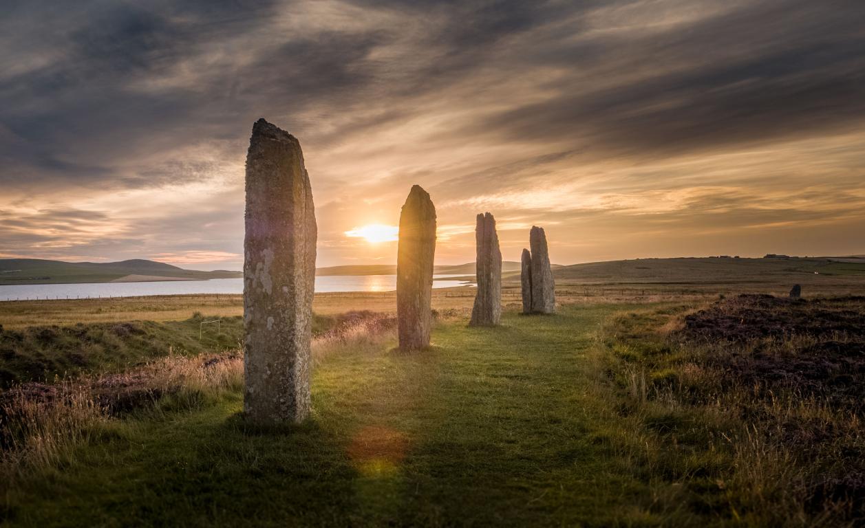 Four large standing stones in a row at the Ring of Brodgar. Dark clouds strewn the sky above the stones as the sun sets behind the stones.