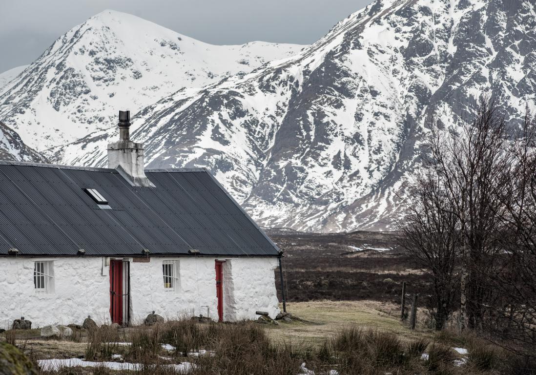 Black Rock Cottage, Glencoe, in winter. The stone cottage building is painted white, with a grey roof, and a narrow red door.