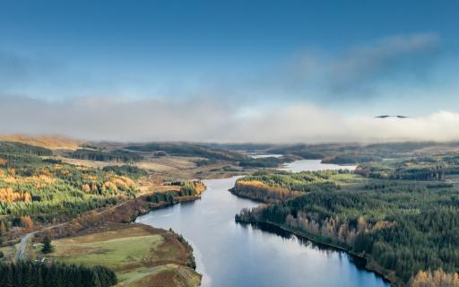 A river runs winds through the centre of the image. The land on both sides of the river is covered in tall green and yellow leaved trees. A layer of mist runs across the length of the image.