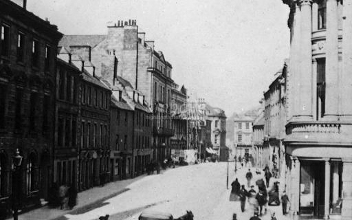 Inverness High Street in 1870. A small dog is seen in the centre of the High Street, along with a horse and carriage. People walk down the pavement on the right hand side of the image.