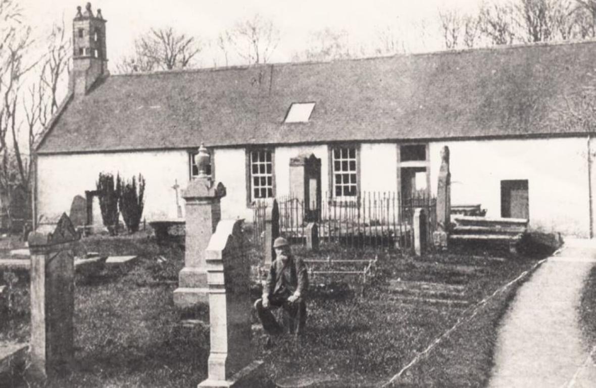 Black and white photograph of a churchyard with tall gravestones. A man wearing a flat cap is crouching down in the centre of the image.