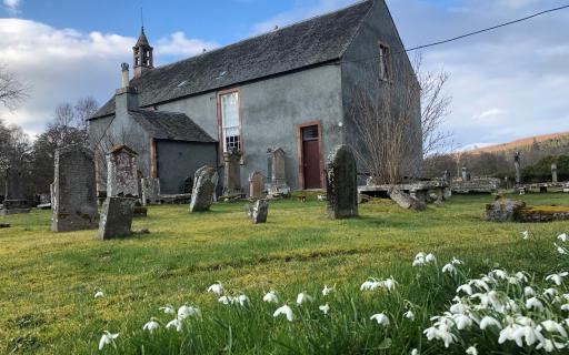 A grey church building with large gravestones in the foreground. Snowdrops can be seen in the kirkyard in the bottom 25% of the image