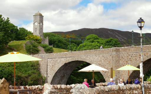 Arched bridge goes left to right of image with outdoor eating area in foreground and mountain backdrop.