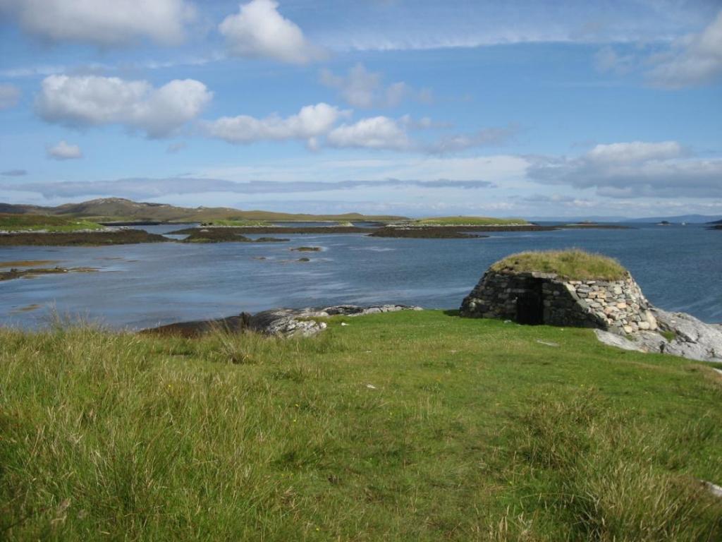 The Hut of Shadows, a small stone circular building with turf on its roof, situated on the right hand side of the image. The hut sits on grassy headland with the sea below.