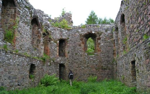 The ruined walls of a stone castle. A person wearing a black jacket and light trousers has their back to the camera in the back, center of the image