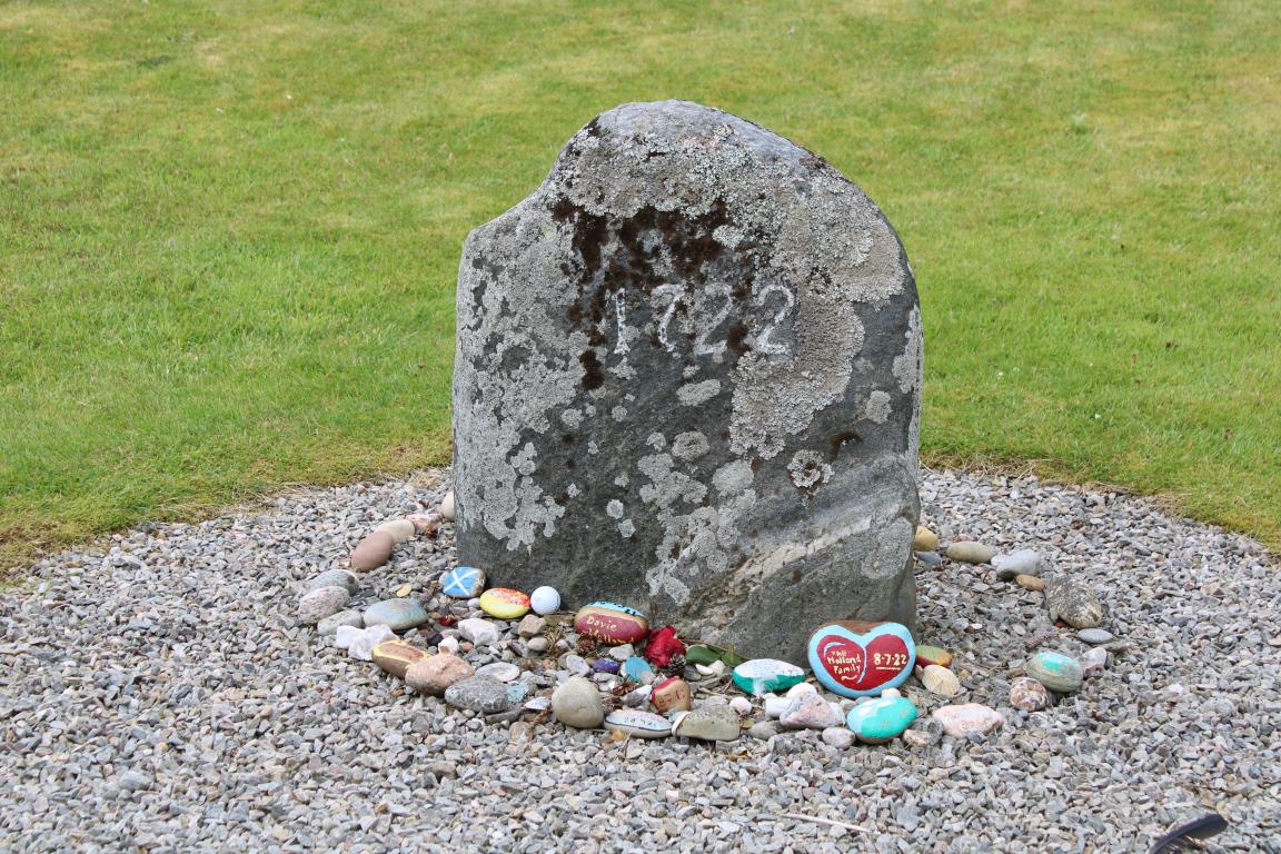 A grey stone with the year '1722' marked in white on its surface. Around the stone is a ring of small stones, both painted and natural, left in tribute.