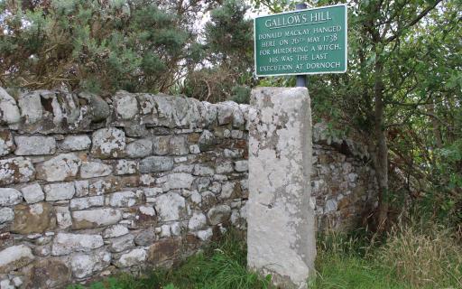 A large grey stone pillar stands in grass next to a large stone wall. Above the pillar, a green sign with white lettering reads 'Gallow's Hill. Donald Mackay hanged here on 26th May 1738 for murdering a witch. His was the last execution at Dornoch'.