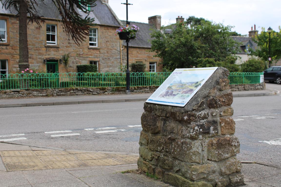 A stone pillar with an interpretation board stands on the side of a road. In the background, two brown brick buildings with grey roofs line the road. A green fence stands in front of these buildings.
