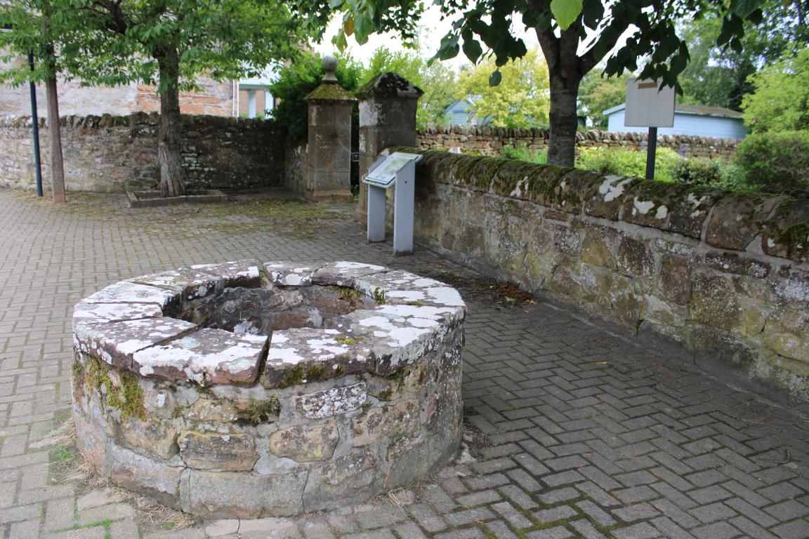 A large stone well stands as the focus of the image. A grey interpretation board is visible in the background, standing adjacent to a stone wall.