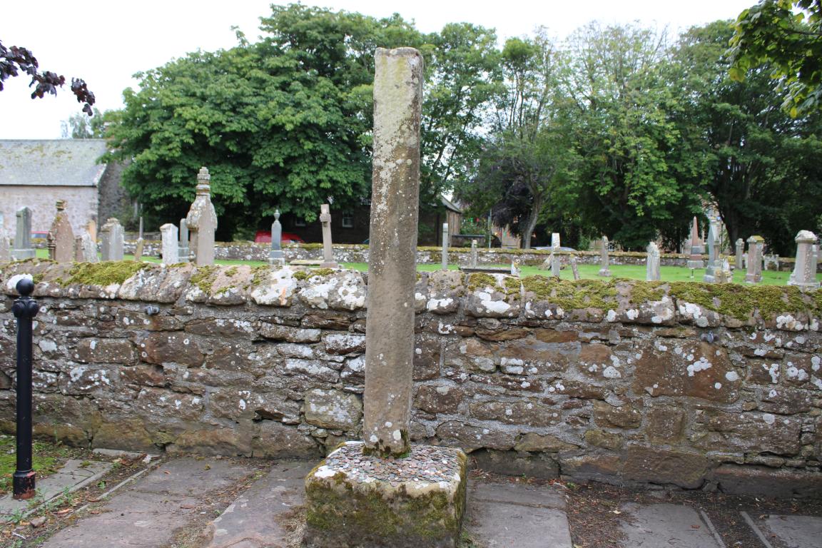 A large grey stone pillar stands on a stone base in the centre of the image. A stone wall stands in the background, with gravestones and trees visible over the wall.